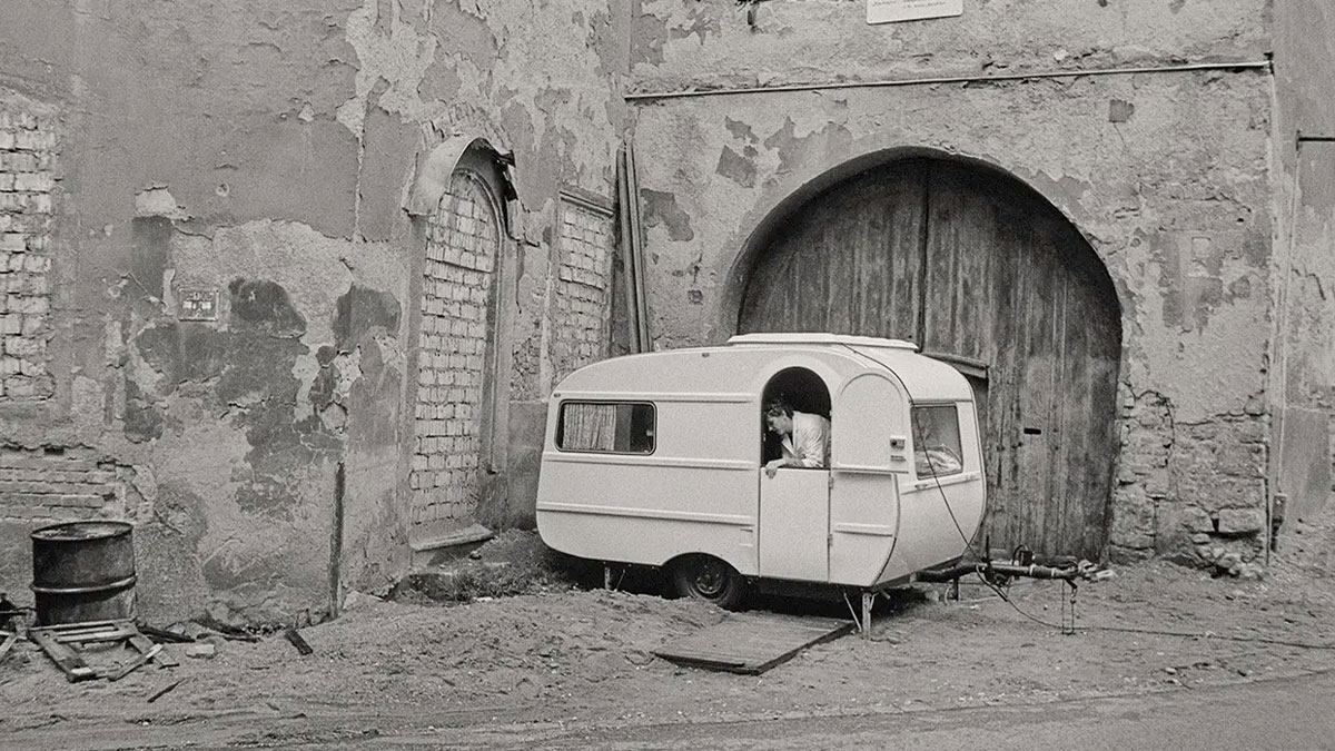 Old East German street scene with a man looking out from a small caravan near a worn, textured wall.