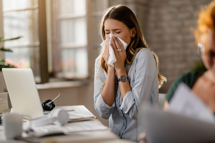 Woman at desk sneezing into tissue while working on laptop, highlighting the fading use of thank you notes in modern settings.