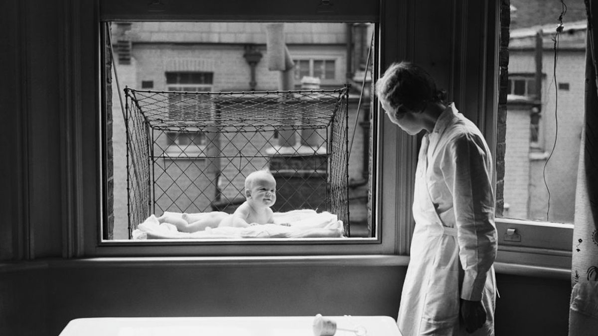 Black and white photo of a baby in a netted balcony crib, an unusual invention for safe outdoor infant care.