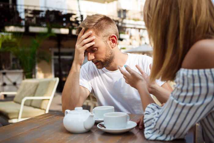 A stressed man holding his head while a woman talks to him at an outdoor café indicating signs of a troubled couple.