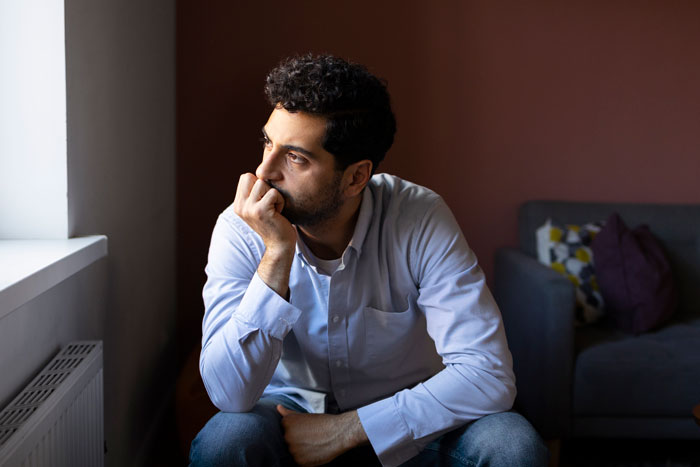 Man looking thoughtful and worried by a window, representing a husband who feels sick after realizing their son’s namesake. Man looking thoughtful and worried by a window, representing a husband who feels sick after realizing their son’s namesake.