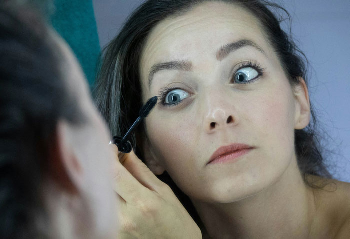 Woman applying mascara to her eyelashes looking into a mirror, enjoying a makeup routine with precise attention. Woman applying mascara to her eyelashes looking into a mirror, enjoying a makeup routine with precise attention.