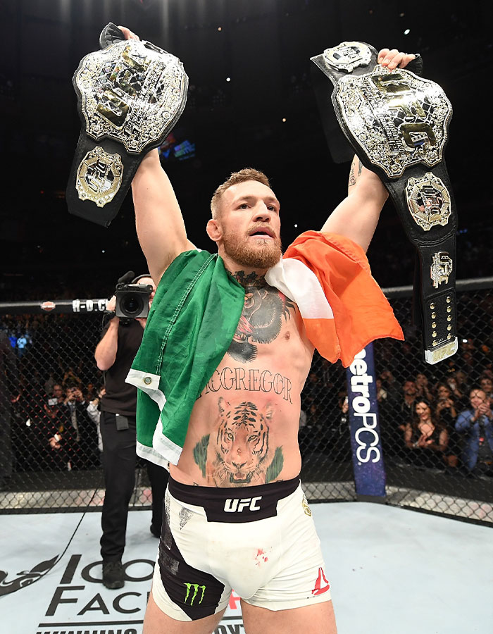 Conor McGregor celebrating victory in UFC octagon, holding two championship belts with Irish flag draped over shoulders. Conor McGregor celebrating victory in UFC octagon, holding two championship belts with Irish flag draped over shoulders.