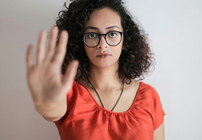 Woman with curly hair and glasses wearing an orange top, holding up her hand as if giving a shade in makeup battle. Woman with curly hair and glasses wearing an orange top, holding up her hand as if giving a shade in makeup battle.