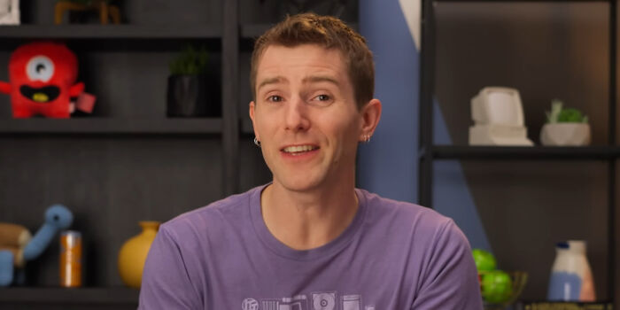 A man in a purple shirt smiling indoors with shelves in the background, illustrating people share unsubscribed YouTubers.