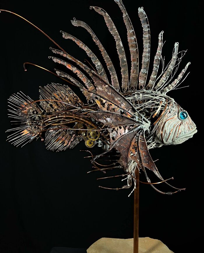 Intricate scrap metal animal sculpture of a fish with detailed fins and bright blue eyes on a stand against black background.