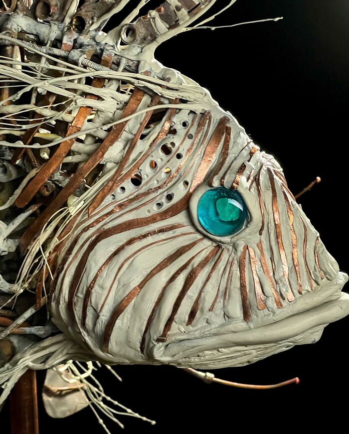 Intricate scrap metal animal sculpture of a fish with detailed fins and bright blue eyes on a stand against black background.