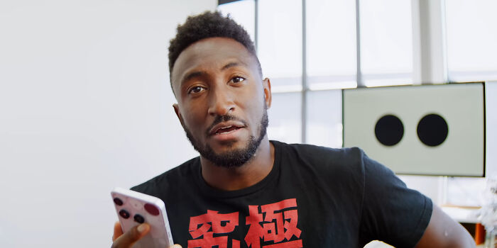 Man holding smartphone speaking about people who share unsubscribed YouTubers in a bright room with windows.