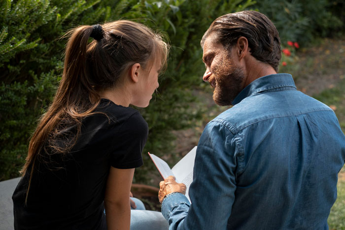 Father and daughter sitting outdoors, sharing a moment while reading a book during a menstruation celebration event. Father and daughter sitting outdoors, sharing a moment while reading a book during a menstruation celebration event.
