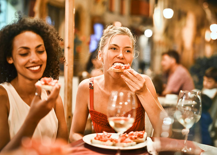 Two women enjoying pizza at a restaurant, reflecting on the bizarre family habits they thought were normal.