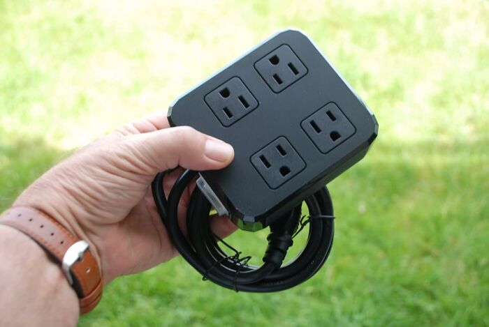 Hand holding an under-the-radar power strip with four outlets and a coiled cord against a grassy background