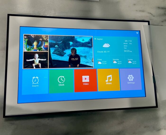 Smart home digital display showing weather, alarm, clock, video, music, and settings apps, highlighting Prime Day electronic deals.