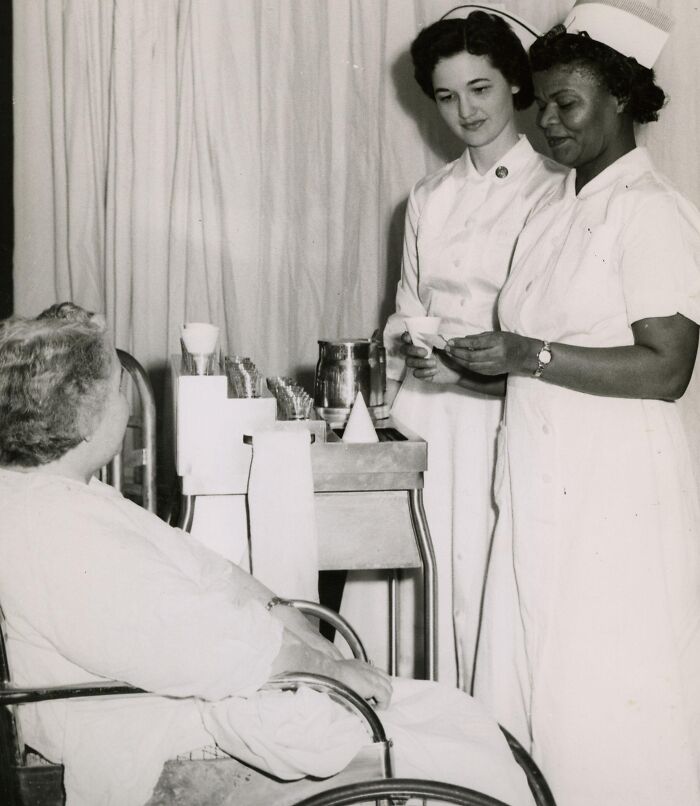 Two vintage nurses in white uniforms attending to a seated patient, showcasing classic nursing care and professionalism.