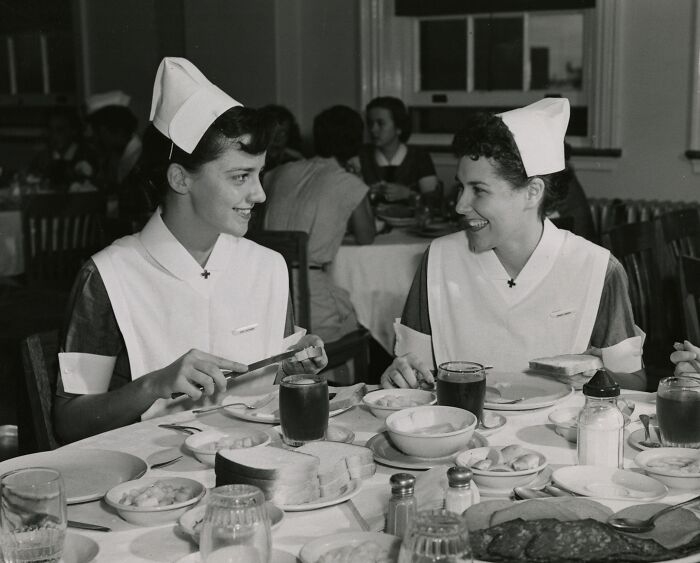 Two vintage nurses in uniform smiling at each other while preparing sandwiches at a dining table.