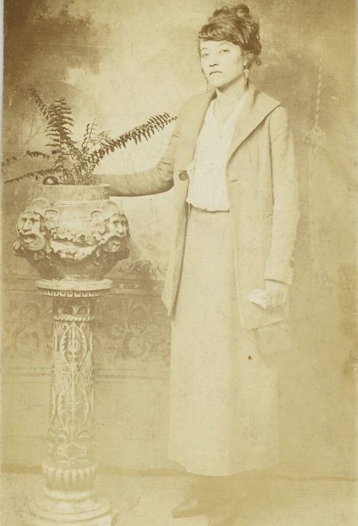 Vintage postcard from the 1930s featuring a woman in period clothing standing next to an ornate plant stand with a fern.