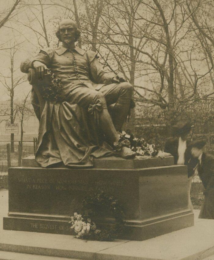 Vintage postcard depicting a 1930s statue with floral wreaths and two blurred people nearby in an outdoor park setting.