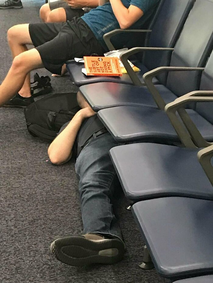 Person lying on the floor under chairs in a public waiting area, showing suspiciously unbothered attitude.
