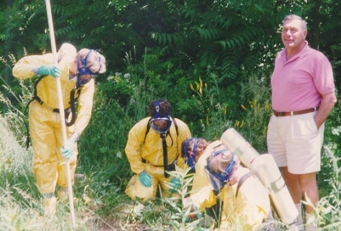 A man in casual clothes stands suspiciously unbothered while others in hazmat suits work in a grassy outdoor area.