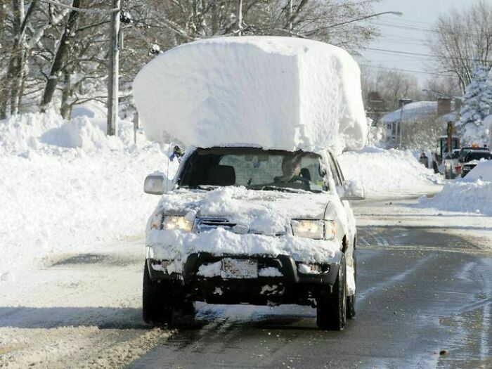 Car driving with suspiciously unbothered attitude carrying large snow pile on roof while roads are snowy and icy.