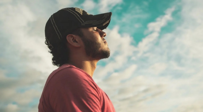 Young man wearing a cap breathing fresh air outdoors with a clear sky, representing gas humans need to breathe to survive.