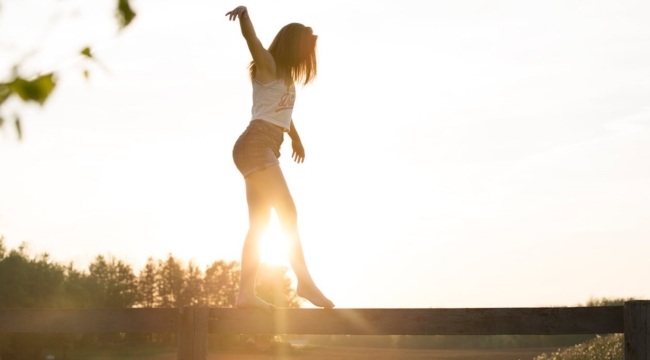 Young woman balancing on a wooden fence outdoors at sunset, illustrating the body part that helps maintain balance.
