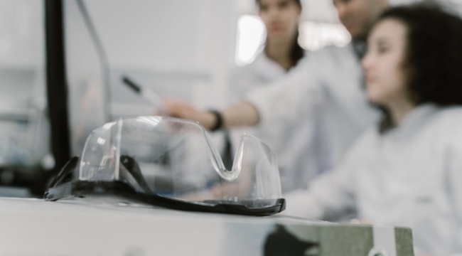 Safety goggles resting on a lab table with scientists in the background studying small objects using scientific tools.