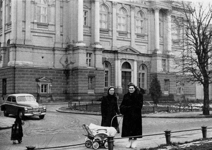 Two women in coats with a stroller on a city street, vintage car and historic building in daily life 50s photo.