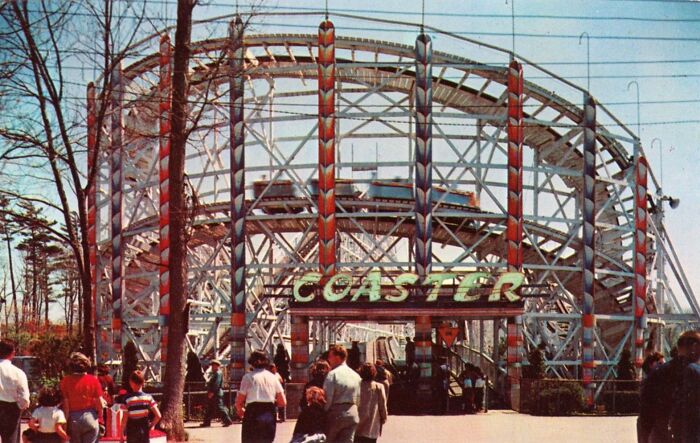 People gathered in front of a classic wooden roller coaster, showcasing daily life and leisure in the 50s.