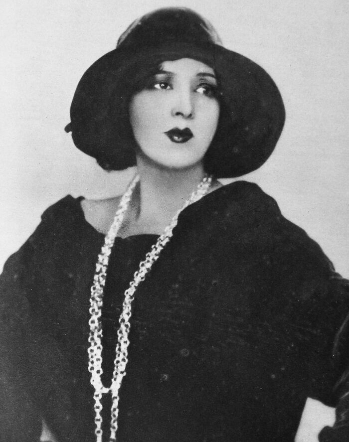 Vintage black and white photo of a woman in a large hat and long necklace, showcasing timeless beauty from a century ago.