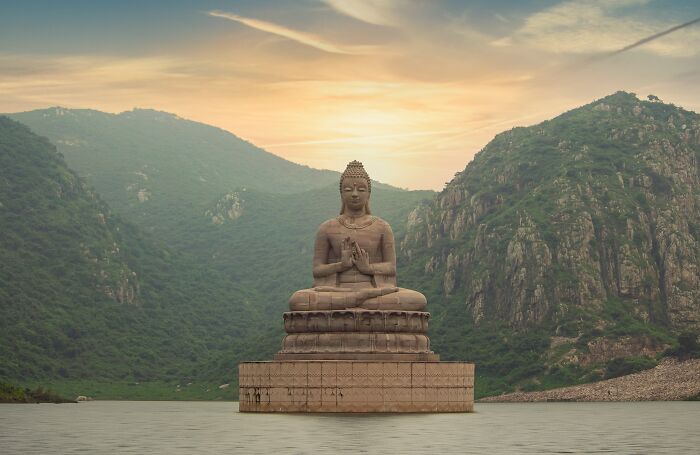 Large stone Buddha statue meditating on a platform in water with green mountains and a sunset sky in the background.