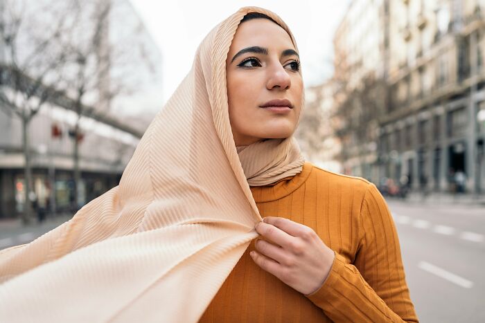 Young woman wearing a beige hijab and orange top standing on a city street, reflecting on Jesus wrote the Bible myths.