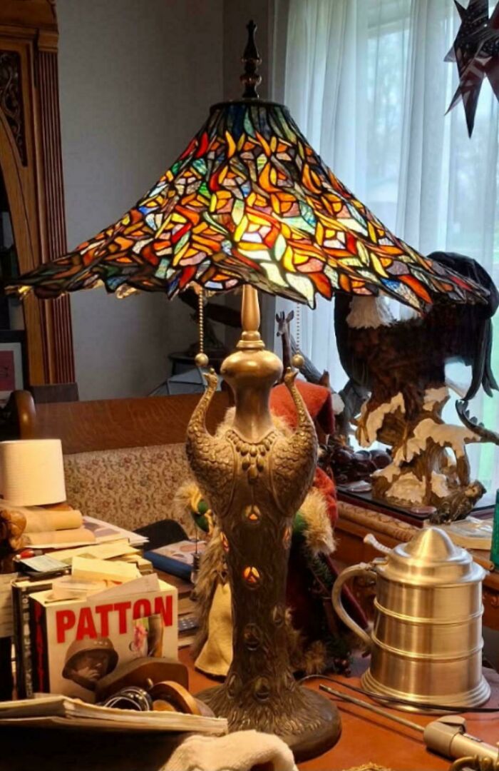 Antique peacock lamp with stained glass shade on a cluttered wooden table among old books and vintage items.