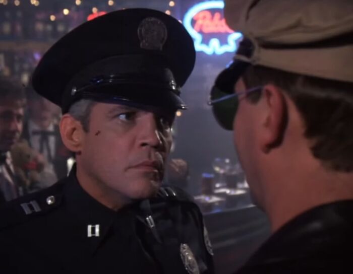 Police officer confronting a man wearing sunglasses in a dimly lit bar scene from cinematic crimes movie sequels.