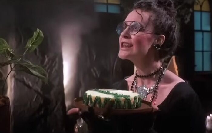 A woman wearing glasses and dark clothing holding a cake in a dimly lit room, cinematic crimes movie scene.