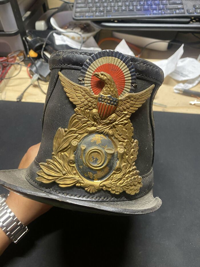 Old vintage military hat with detailed gold eagle emblem and patriotic insignia held indoors on a cluttered desk surface
