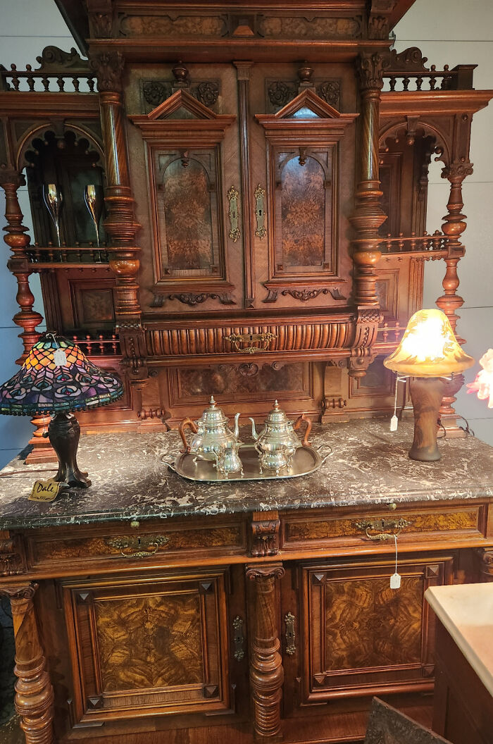 Intricately carved antique wooden cabinet with marble top, vintage silver tea set, and decorative lamps showcasing beautiful old things.