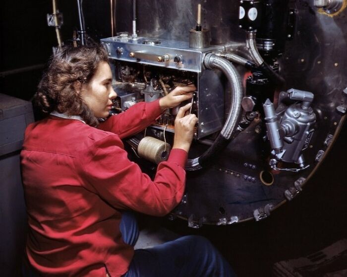 Woman at work during WWII, wearing a red jacket and fixing machinery in an industrial setting.