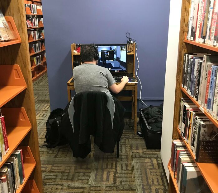 Man sitting unbothered at a computer desk in a library, focused on screen despite quiet surroundings.