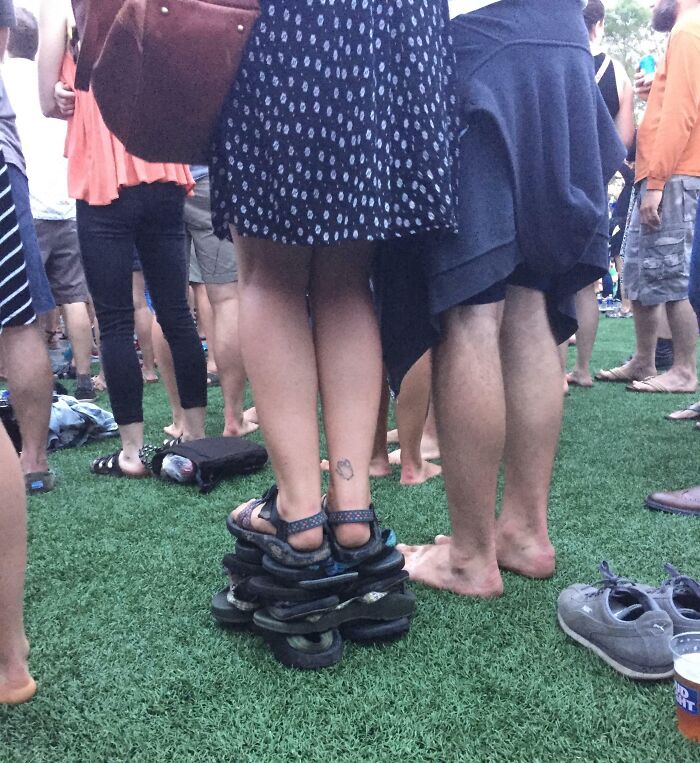 Person standing on stacked sandals at crowded outdoor event, showing suspiciously unbothered attitude amid the crowd.