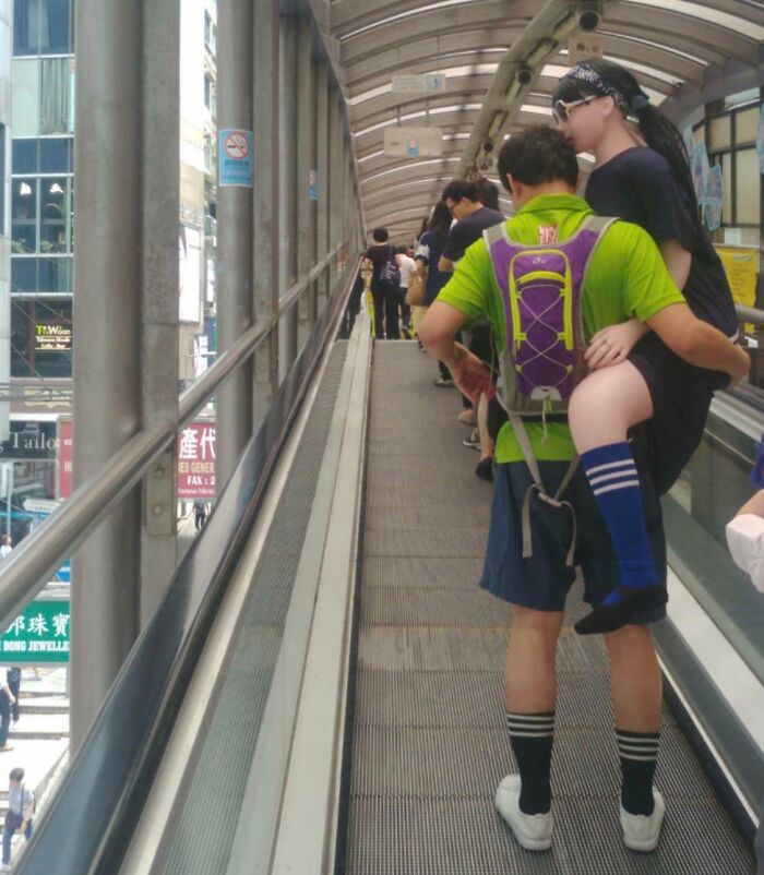 Man wearing a green shirt carries a lifelike doll on a crowded moving walkway, showing suspiciously unbothered behavior.