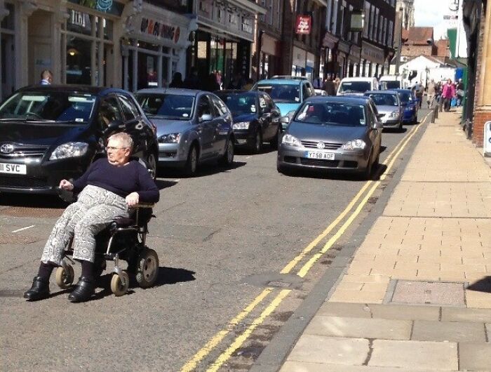 Elderly woman in a wheelchair on a busy street, appearing suspiciously unbothered by the traffic around her.