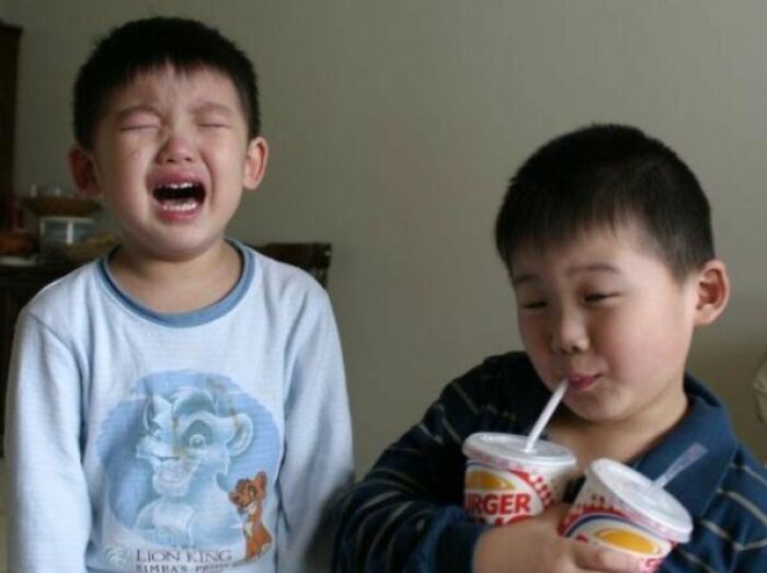 Two young boys indoors, one crying and the other suspiciously unbothered while drinking from fast food cups.