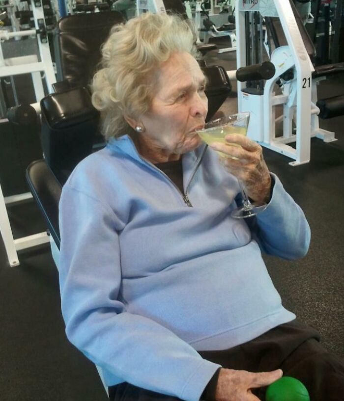 Elderly woman drinking a cocktail at the gym, looking suspiciously unbothered while exercising with weights.