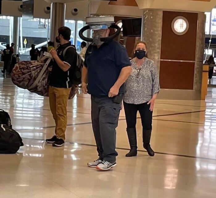 Man at airport wearing unusual suspended face shield and mask, showing suspiciously unbothered attitude amid crowds.