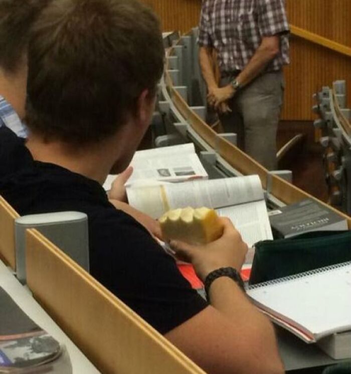Student in lecture hall eating bread while studying, showing suspiciously unbothered attitude during class.