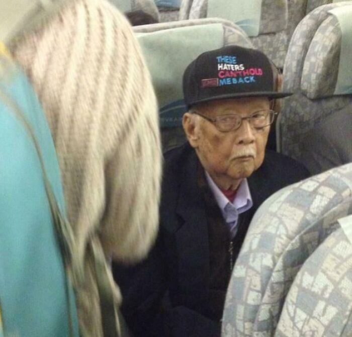 Elderly man wearing a cap with a bold phrase, looking suspiciously unbothered on a crowded bus seat.