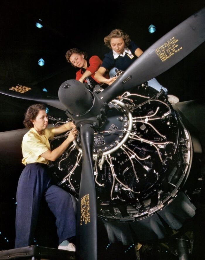 Women at work during WWII repairing a large aircraft engine propeller in a factory setting.