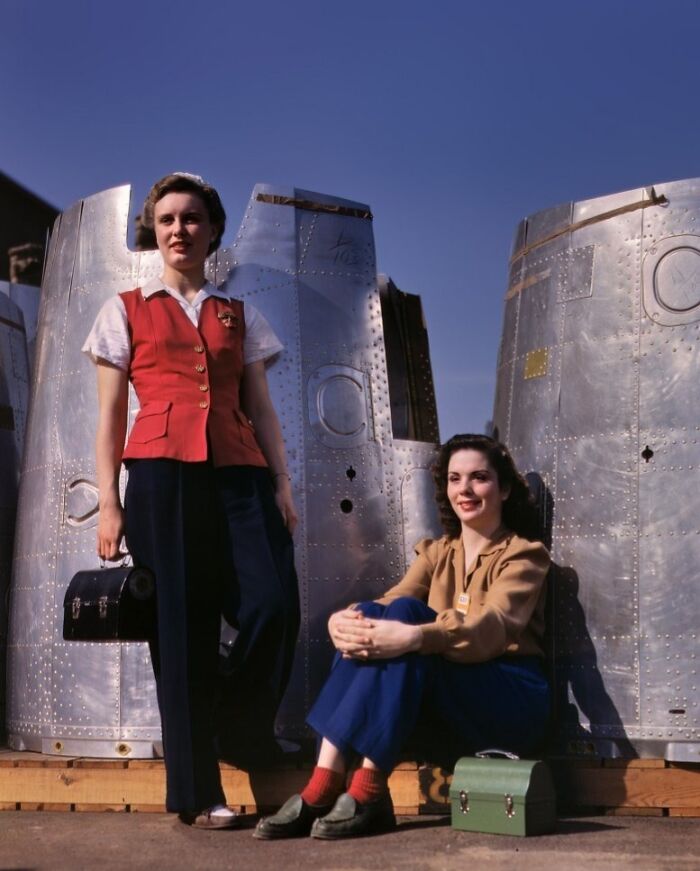 Two women at work during WWII, standing and sitting by metal airplane parts, showcasing women at work in WWII industry.