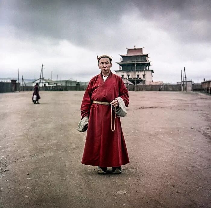 Man in traditional red robe standing on a dusty street with a temple in the background, daily life photo from the 50s.