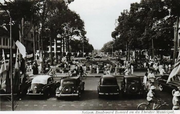 Busy street scene with vintage cars and people on bicycles, showcasing daily life across the globe in the 50s.